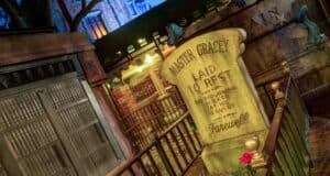 A gravestone inscribed “Master Gracey Laid To Rest” stands in front of an iron gate at night, surrounded by gold fencing and red flowers, with spooky decorations and dim lighting setting an eerie mood.
