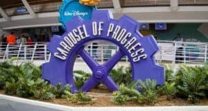 The iconic purple gear-shaped Carousel of Progress sign welcomes guests amid lush greenery at the entrance to Disney World.